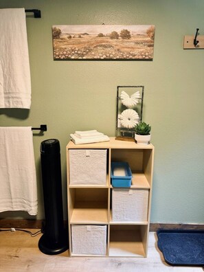 A bathroom with a green wall, a white towel hanging, a black floor lamp, a shelf with folded towels, a framed picture, and a small potted plant.