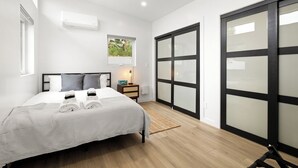 A modern bedroom with a bed, bedside table, lamp, and a view of a garden through a window. - Spacious w/1BR 1Bath - Near DTLA (Los Angeles)