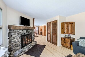A living room with a stone fireplace, wooden cabinets, a flat-screen TV, and a sofa. - Spacious Upstairs Apartment Near Stowe Mountain (Stowe)