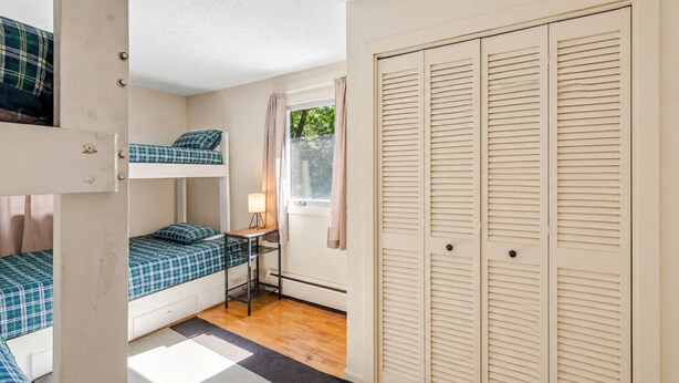 Une chambre avec des lits superposés, une petite table avec une lampe, une fenêtre avec des rideaux et un ensemble de portes à persiennes.