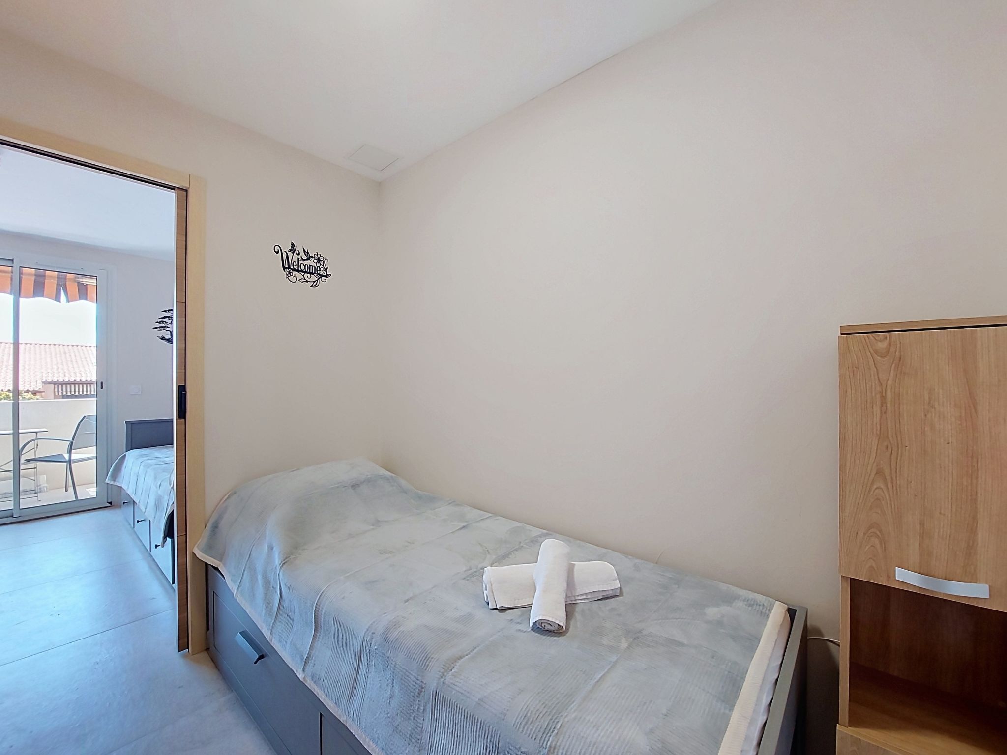 A single bed with a white mattress and a towel, a wooden wardrobe, and a 'Welcome' sign on the wall.
