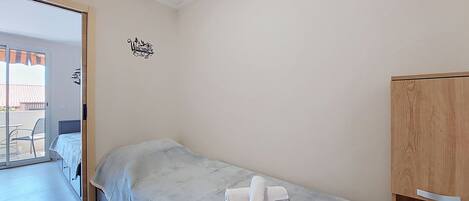 A single bed with a white mattress and a towel, a wooden wardrobe, and a 'Welcome' sign on the wall.