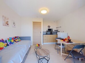 A compact room with a bed, a small dining table with chairs, a kitchenette, and a ceiling light. - Les Aigues Marines by Interhome (Saint Cyr sur Mer La Madrague)