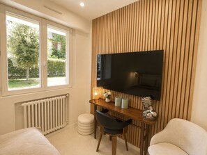 Een moderne slaapkamer met een groot raam, een flatscreen televisie bevestigd aan een houten wandpaneel, een nachtkastje met een lamp en twee stoelen.