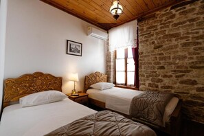 Habitació amb dos llits, sostre de fusta, paret de pedra, finestra amb cortines i aire condicionat fixat a la paret.