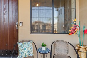 A room with a wooden door, a window with a view of buildings, two wicker chairs, a small table with a plant, and a decorative vase with flowers. - Casa La Quinta ~ Luxury Condo w/Pool near Old Town (La Quinta)