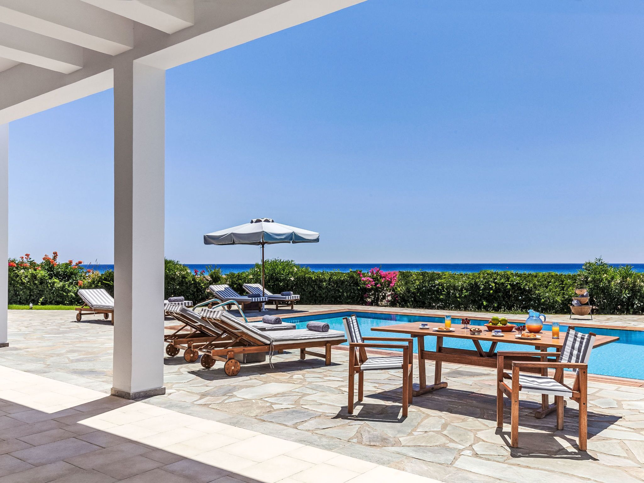 A poolside area with lounge chairs and a table, overlooking a clear blue sea.