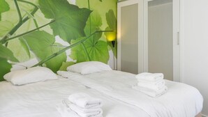 Hotellihuone, jossa on sänky, valkoiset vuodevaatteet, vihreälehtinen seinämaalaus, vaatekaappi ja seinälle asennettu valaisin.