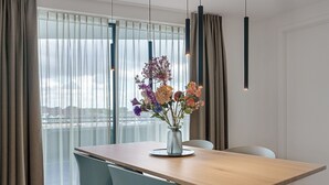 A modern dining area with a wooden table, two chairs, a vase with flowers, and a view of a cityscape through large windows. - 4-guest Apartment in Breskens (Breskens)