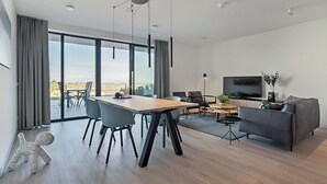 A modern living room with a dining area, a sofa, a TV, and a view through large windows. - Coastal Apartment Stay in Breskens (Breskens)