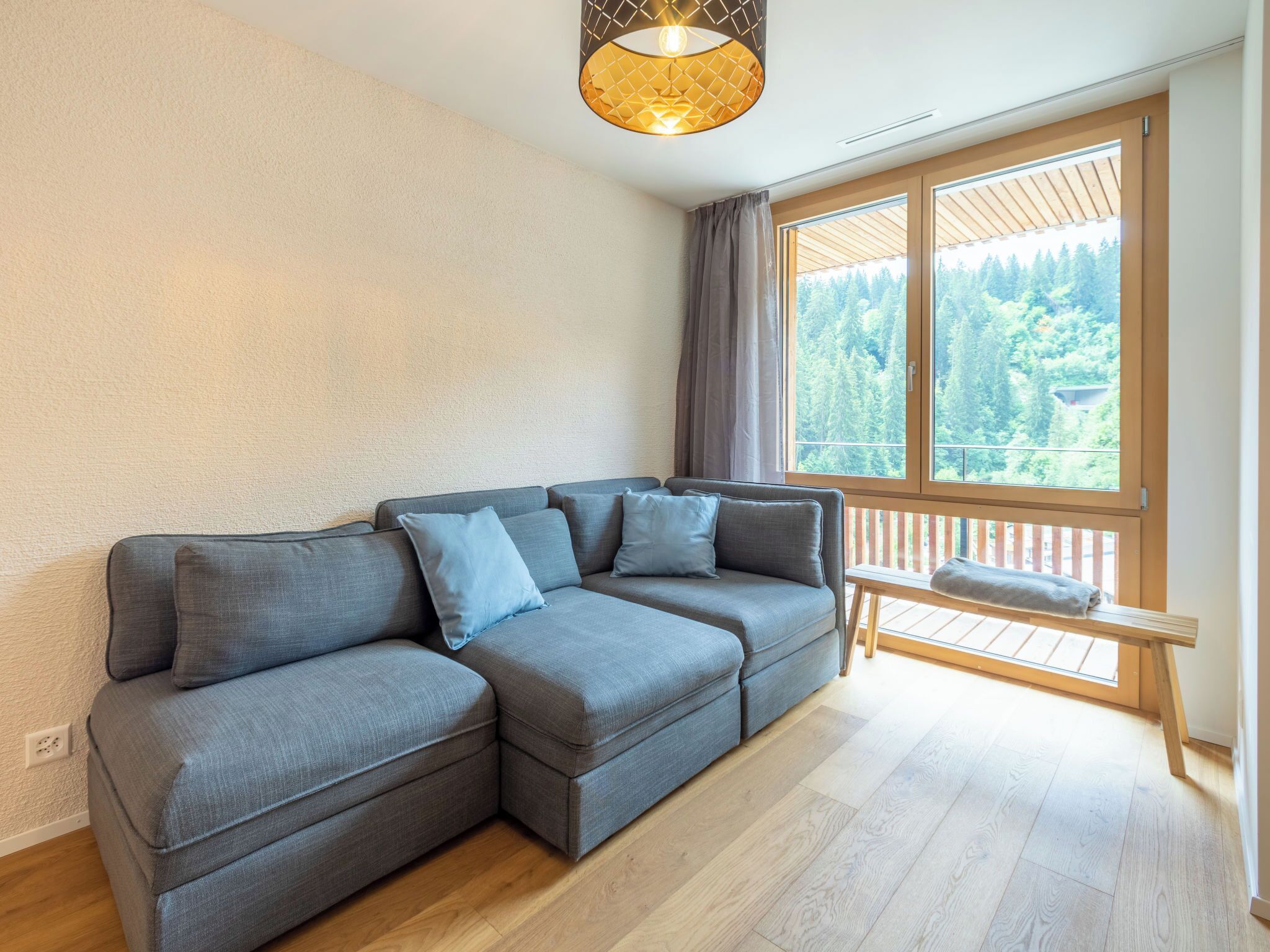 A modern living room with a sectional sofa, a bench, and a large window offering a view of a forest.