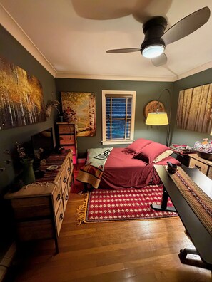 Ein Schlafzimmer mit einem Bett, einem Schreibtisch, einem Stuhl, einer Stehlampe, einem Deckenventilator und Kunstwerken an den Wänden.