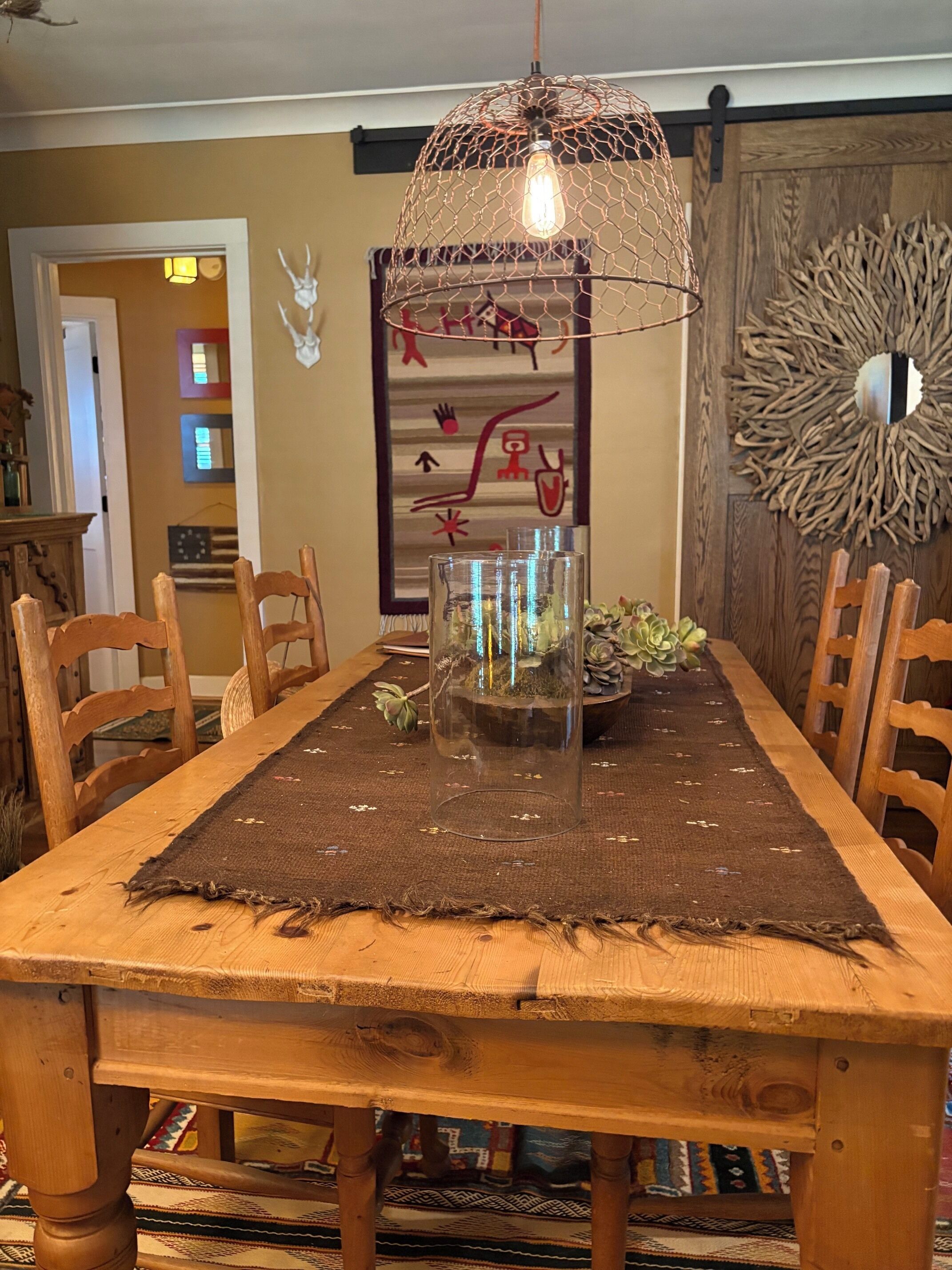 A wooden dining table with chairs, a hanging light fixture, and a wall with decorative items.