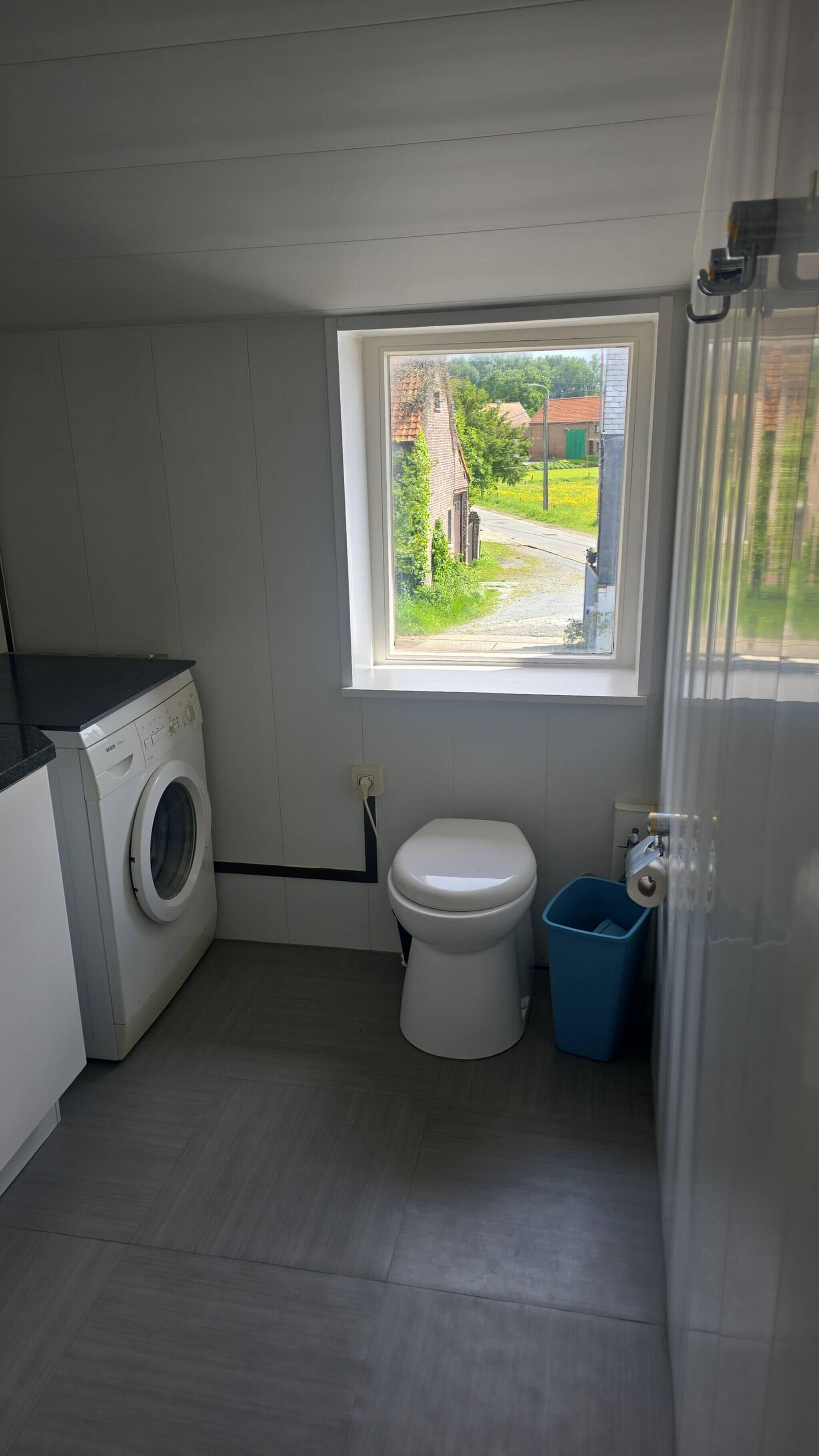 Een compacte badkamer met een wasmachine, toilet en een raam met uitzicht op een landelijk landschap.