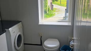 Een compacte badkamer met een wasmachine, toilet en een raam met uitzicht op een landelijk landschap.