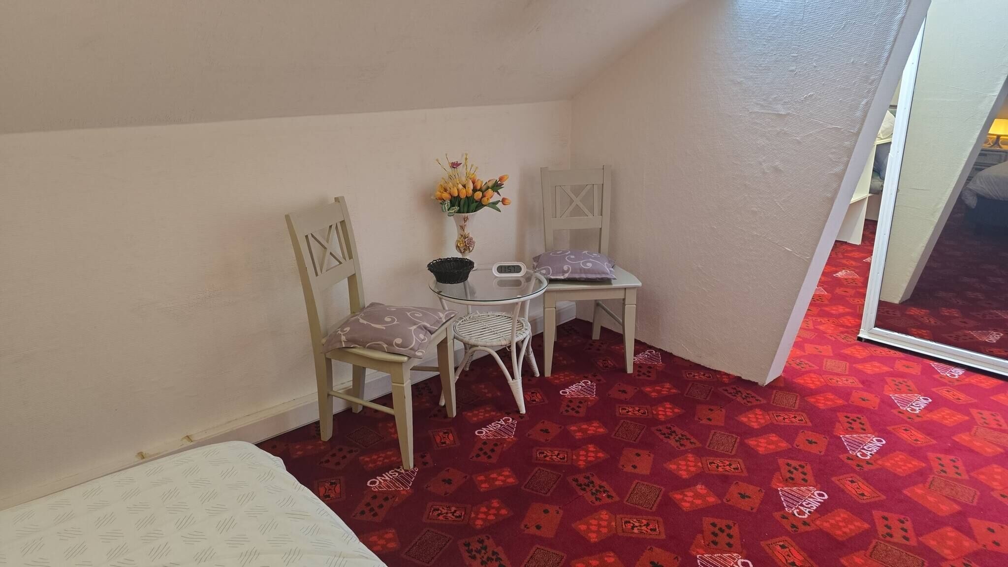 A small room with a bed, a table with chairs, a vase with flowers, and a red patterned carpet.