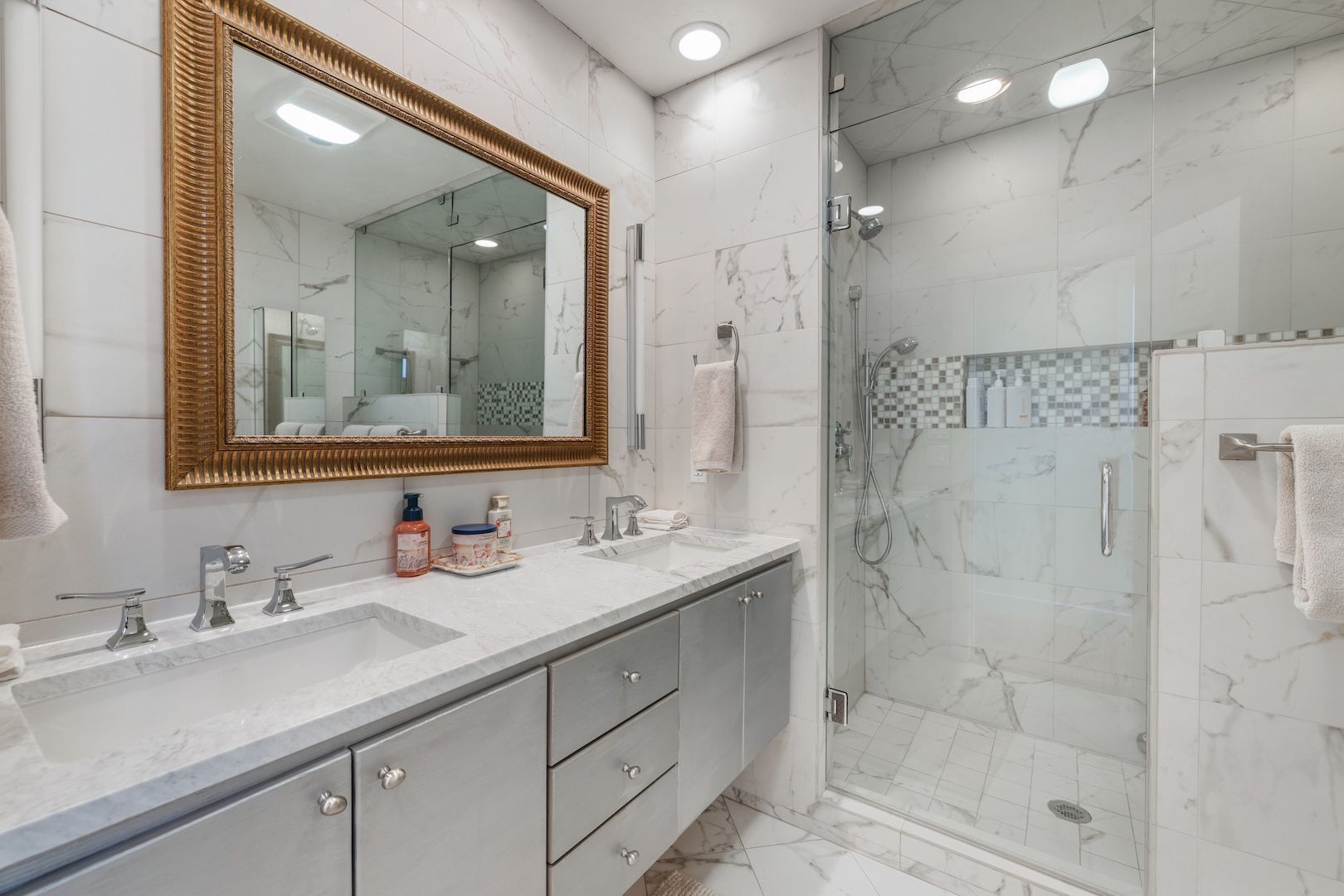 A modern bathroom with a large mirror, two sinks, a shower enclosure, and marble flooring.