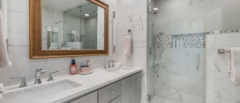A modern bathroom with a large mirror, two sinks, a shower enclosure, and marble flooring.