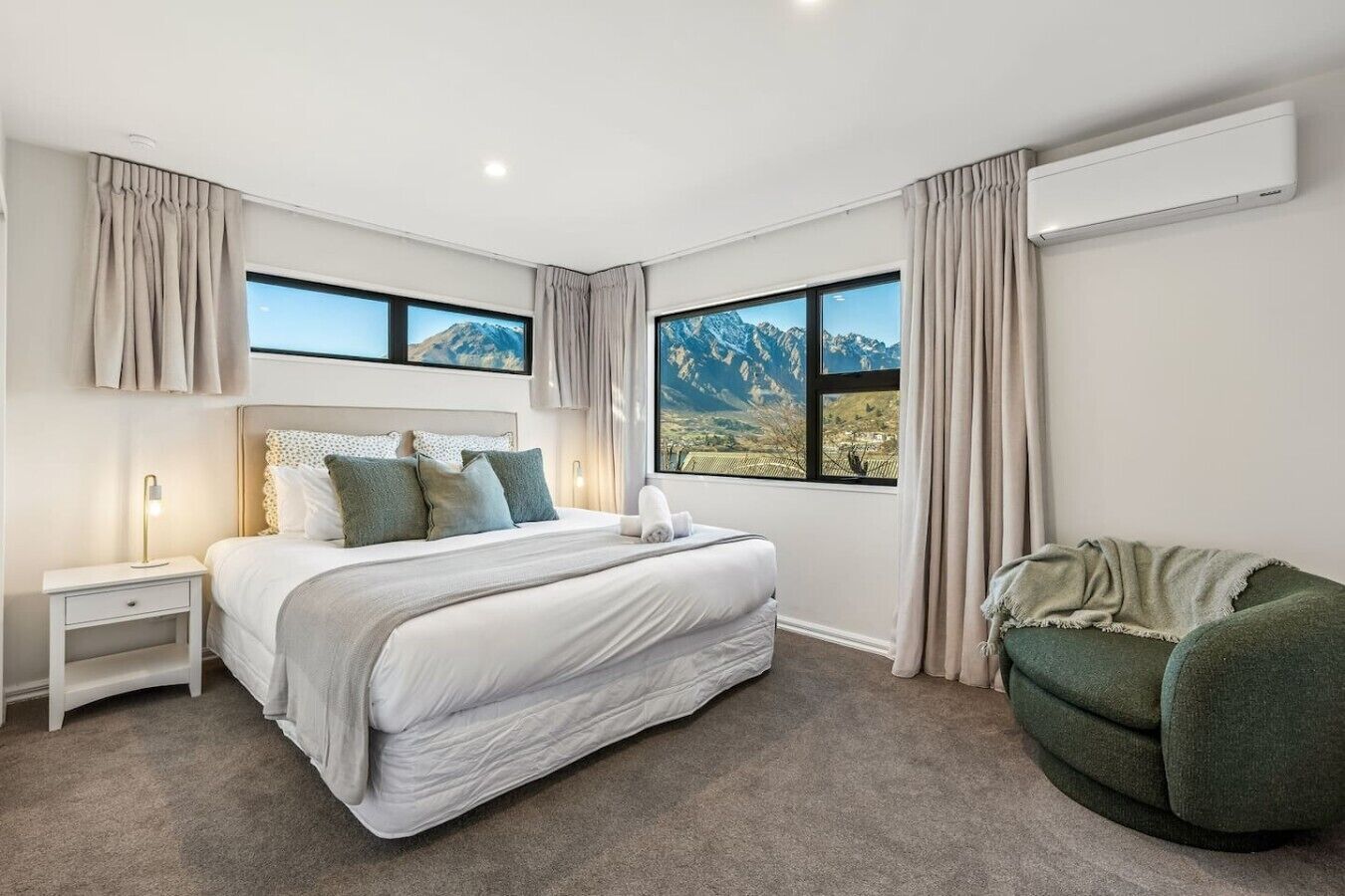 Ein Schlafzimmer mit einem großen Bett, zwei Fenstern mit Blick auf die Berge, einem Nachttisch mit einer Lampe und einem grünen Sessel.