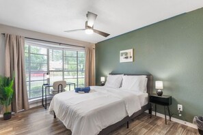 A hotel room with a bed, a desk, a chair, a ceiling fan, and a view through the window. - 2 Min AT&T Stadium Walk | Cozy & Clean 2BR Apt (Arlington)