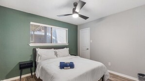 Ein Schlafzimmer mit einem Bett, einem Nachttisch, einem Deckenventilator, einem Fenster mit Jalousien und einer Tür.