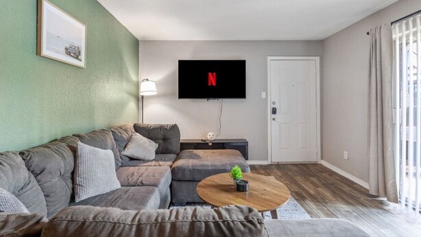Ein Wohnzimmer mit Ecksofa, Couchtisch, flachbildfernseher an der Wand und einer Tür.