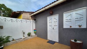Una casa con fachada gris, una puerta y una ventana blancas. Cuenta con un pequeño jardín con plantas y una maceta grande.