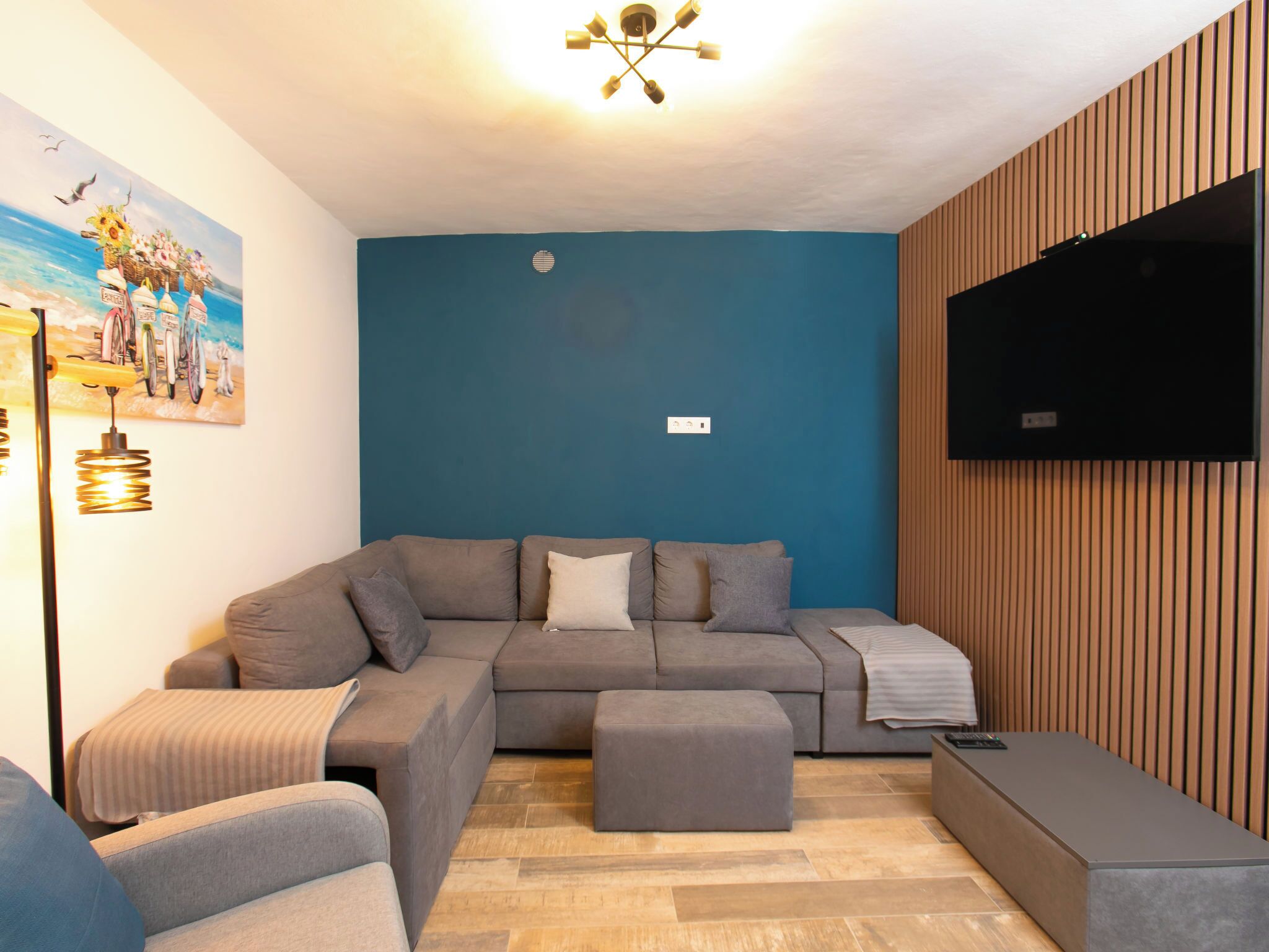 Un salon moderne avec un canapé d’angle, un téléviseur à écran plat fixé sur un mur en bois et un tableau représentant des bicyclettes.