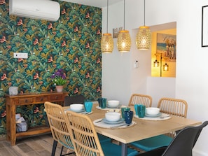 Uma área de refeições com papel de parede tropical, cadeiras de vime e uma mesa de madeira posta com pratos azuis e brancos.