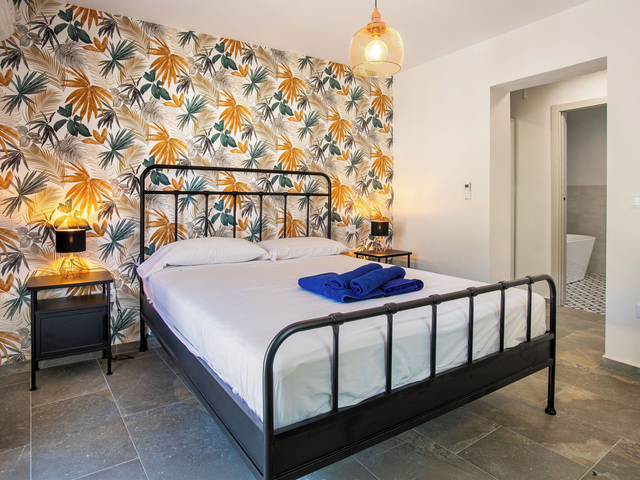 Une chambre à coucher avec un lit, deux lampes de chevet, un mur orné d’un motif de feuilles tropicales et une porte donnant sur une autre pièce.