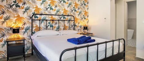 Un dormitorio con una cama, dos lámparas de noche, una pared con un diseño de hojas tropicales y un umbral que conduce a otra habitación.