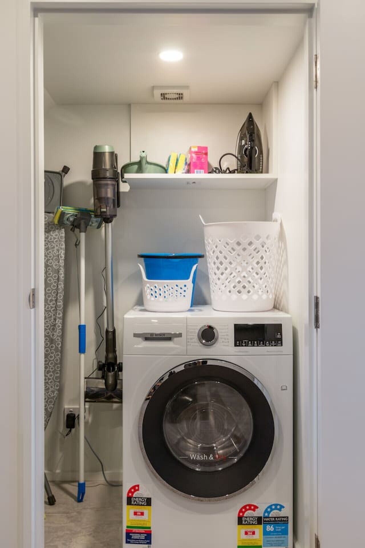 A laundry room with a Bosch washing machine, a drying rack, and a shelf with various items.