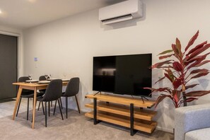 Ein modernes Wohnzimmer mit einem Flachbildfernseher auf einem Holzboden, einem Esstisch mit Stühlen und einer großen Topfpflanze.
