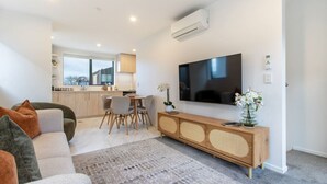 Una sala moderna con sofá, una mesa de comedor con sillas, un televisor de pantalla plana montado en la pared y una cocina al fondo.