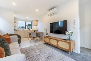 Ein modernes Wohnzimmer mit einer Couch, einem Esstisch mit Stühlen, einem flachbildfernseher an der Wand und einer Küche im Hintergrund.