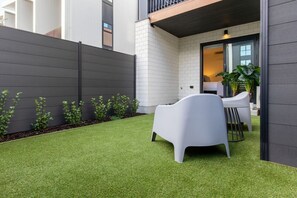 Un espace extérieur moderne aménagé avec des sièges, comprenant une chaise et une table blanches, un petit jardin avec du gazon synthétique, et un bâtiment à l’extérieur sombre et aux murs en briques blanches.