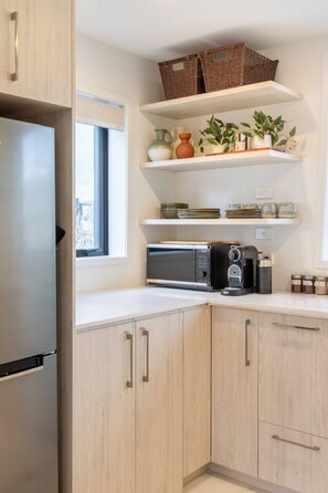 Eine Küche mit hellen Holzschränken, einem Edelstahlkühlschrank, einer Mikrowelle und einer Kaffeemaschine. Über der Arbeitsplatte befinden sich Regale mit Körben und Küchenutensilien.