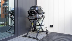 Una parrilla portátil con tanque de gas y tapa, ubicada sobre un soporte con ruedas, al lado de un edificio con ventana y puerta.