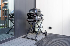 Ein tragbarer Grill mit Gasflasche und Deckel, auf einem Rollgestell stehend, neben einem Gebäude mit Fenster und Tür.