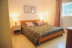 A bedroom with a wooden bed, two bedside tables with lamps, a nightstand, and a window with curtains.