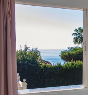 Blick aus einem Fenster mit Vorhang auf eine Küstenlandschaft mit Grünflächen und blauem Himmel.