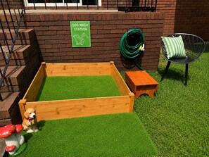 A dog wash station sign, a wooden dog bed with artificial grass, a green garden hose, and a chair with a striped cushion. - Dog Lovers' Dream  Wash Station + Dog Library (Warner Robins)