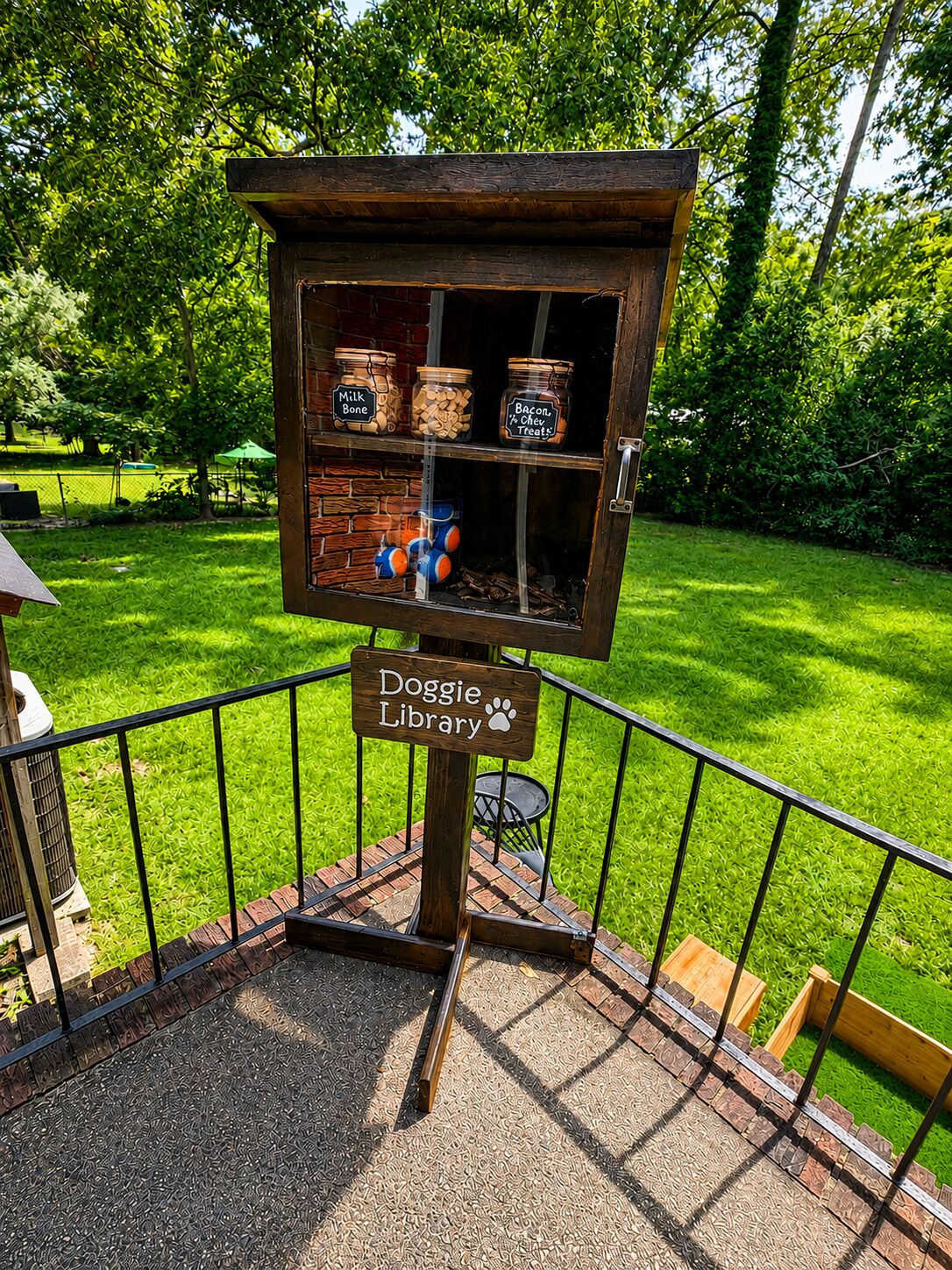 A Doggie Library with a brick base and metal railing, containing jars labeled 'Milk Bone,' 'Bacon & Chew Treats,' and 'Bacon.'