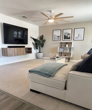 Ein modernes Wohnzimmer mit Flachbildfernseher, Deckenventilator, einer großen Topfpflanze und einem Sofa mit Decke.