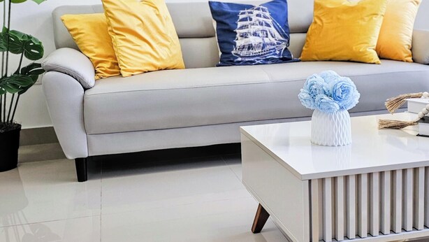 Ein modernes Wohnzimmer mit einem grauen Sofa, gelb-blauen Kissen, einem weißen Couchtisch und zwei gerahmten floralen Bildern an der Wand.