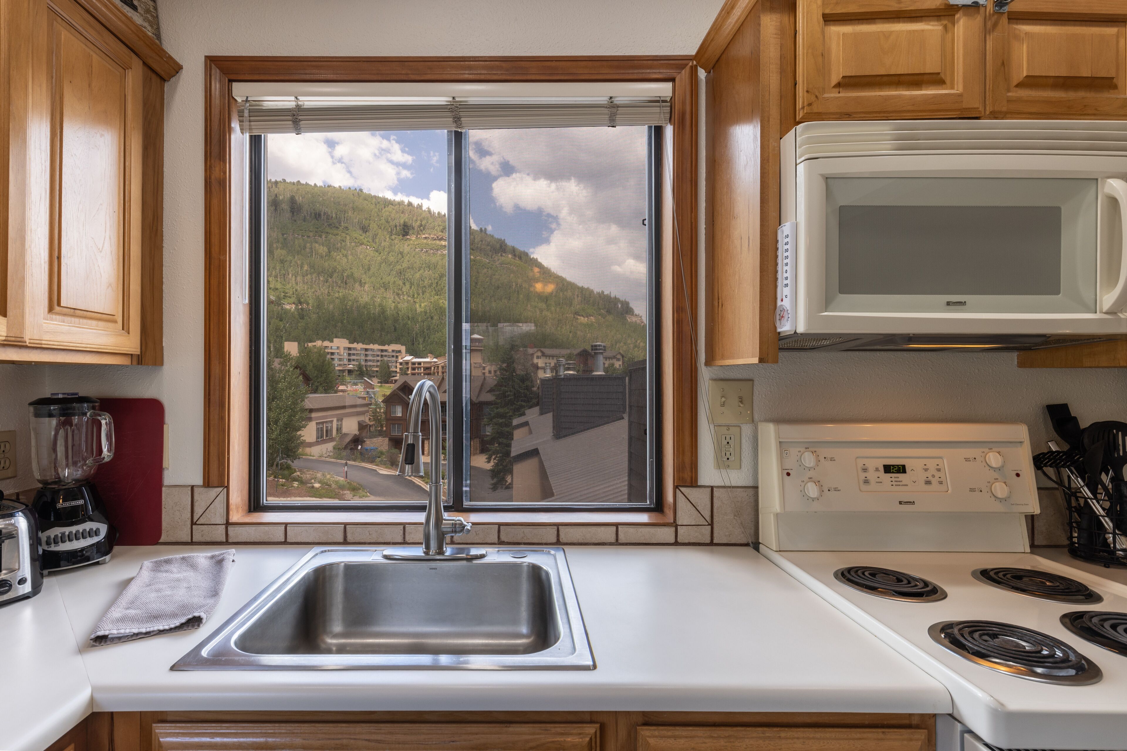 A kitchen with a stainless steel sink, a white stove, and a window with a view of a hilly landscape.
