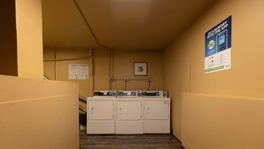Een wasserette met een rij wasmachines en drogers, een poster die een mobiele app promoot, en een ingelijst beeld aan de muur.