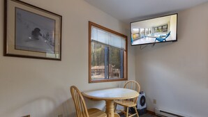 Een kamer met een eettafel en stoelen, een wandtelevisie, een raam met zonwering en een foto in een lijst aan de muur.