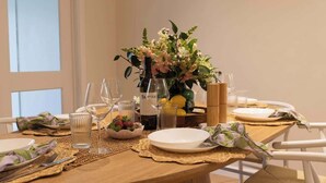 Ein Esstisch ist gedeckt mit Tellern, Besteck, Gläsern und einer Weinflasche, dazu stehen eine Blumenvase und eine Obstschale auf dem Tisch.