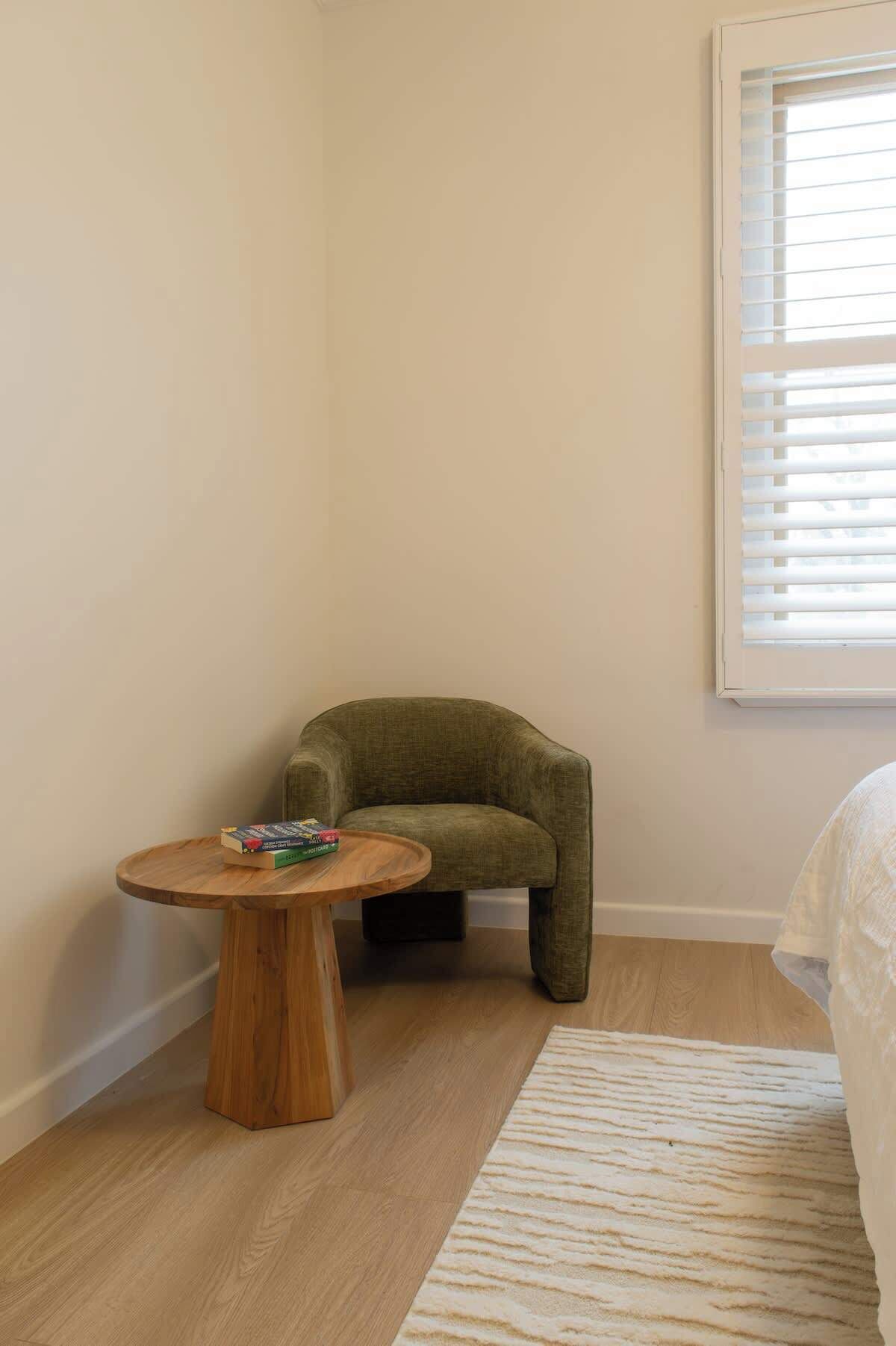 A small room with a round wooden table, a green armchair, a white bed, and a window with blinds.